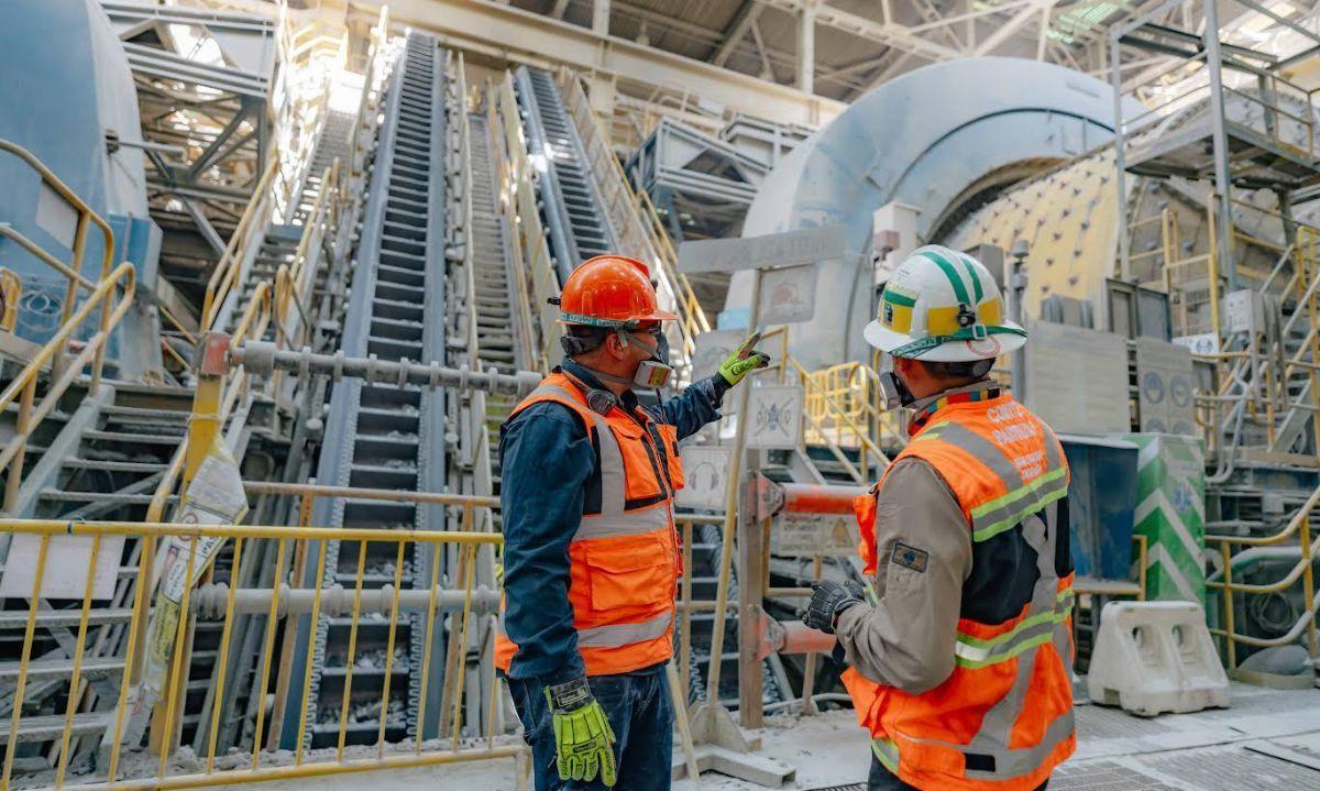 Trabajadores con casco y chaleco reflectante observando una planta de procesamiento minero al atardecer